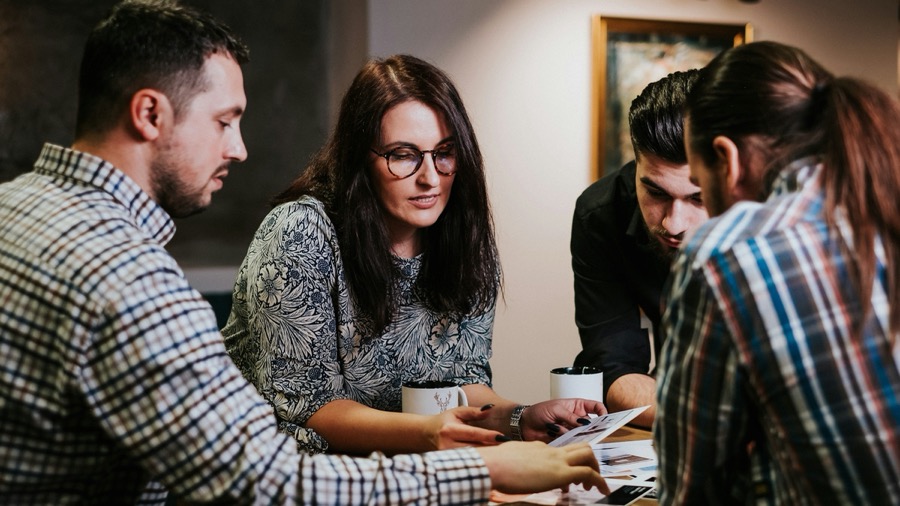 Four people gathered close around one end of a wooden table, leaning in over printed pages and coffee cups while one of them points something out.